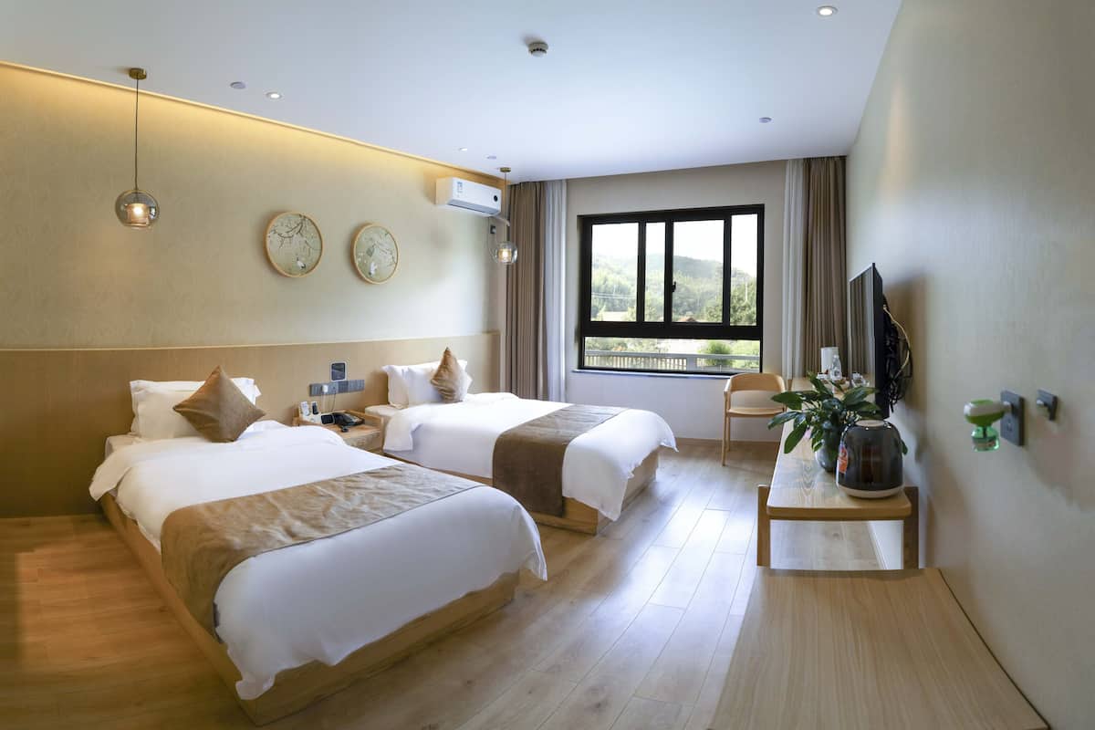 A hotel room with two beds, a wooden floor, a TV, a small table with a plant, and a window with a view.