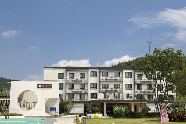 A multi-story hotel with a modern design, featuring balconies with plants, a large circular window on the ground floor, and a windmill sculpture in the foreground. - Paradise hotspring hotel (Chenzhou)