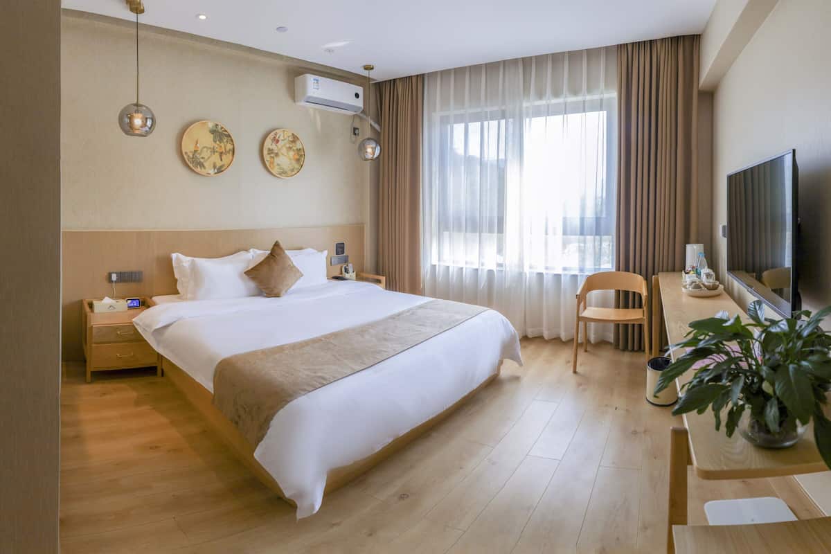 A hotel room with a large bed, bedside tables, a desk with a chair, a television, and a potted plant.