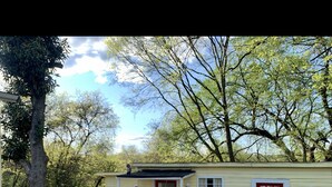 A small, single-story building with a red door and windows, surrounded by trees and a gravel path. - Charming Country Cottage- Long Term Stays Welcome! (Eden)