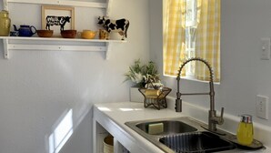 A compact kitchen with white countertops, a stainless steel sink, and a window with yellow curtains. - Charming Country Cottage- Long Term Stays Welcome! (Eden)