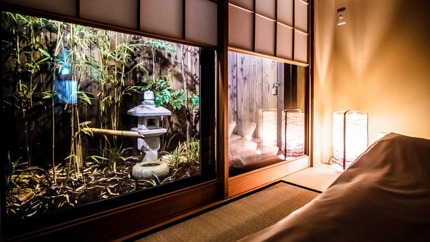 Huone, jossa on perinteinen japanilaistyylinen liukuovi, pieni sisäpuutarha, jossa on bambua, kivinen suihkulähde ja valkoisella peitteellä varustettu sänky.