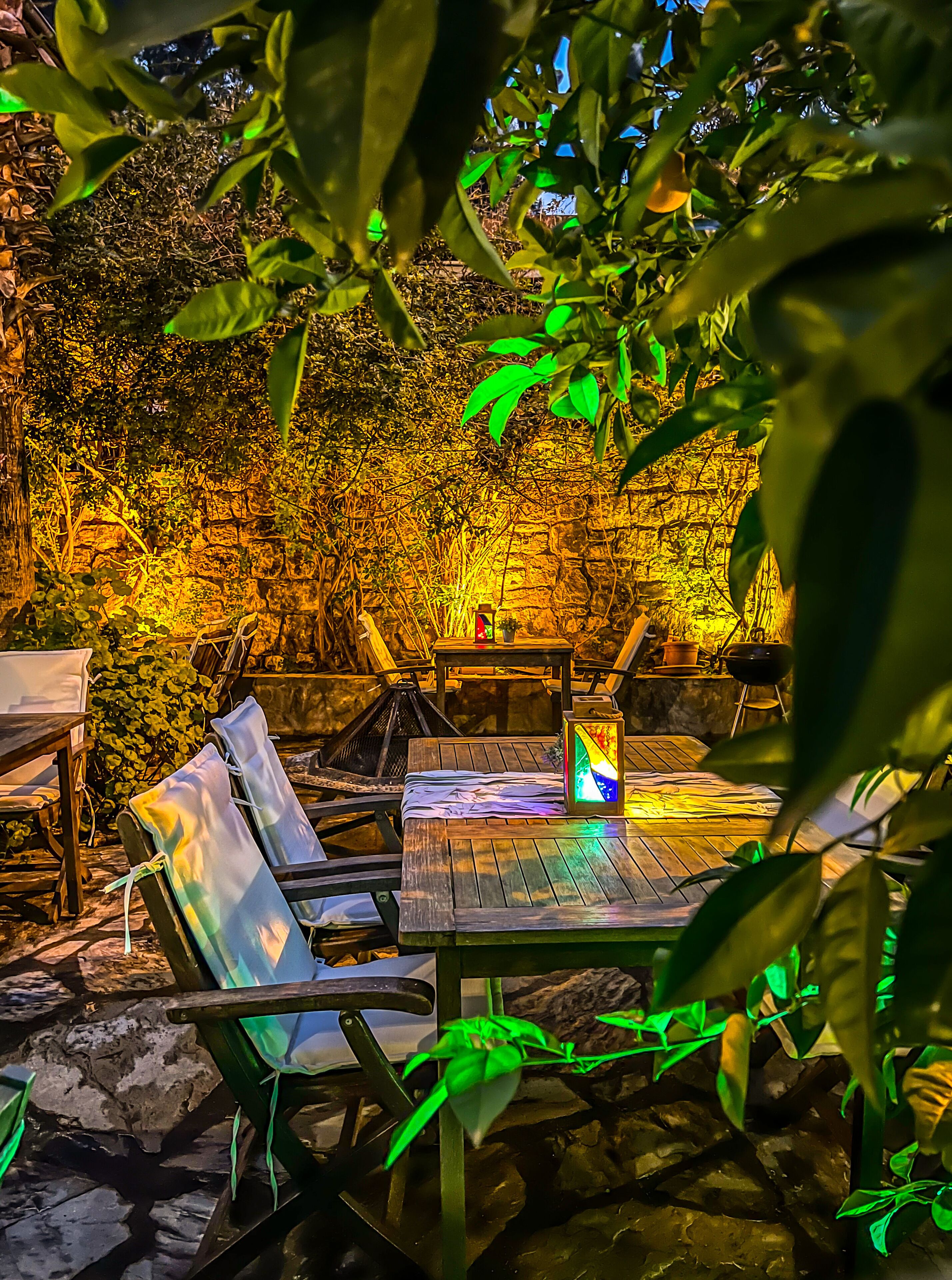 An outdoor seating area with a table and chairs, surrounded by lush greenery and a stone wall.