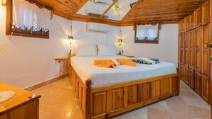 Una cama con estructura de madera, un colchón blanco, dos almohadas y una toalla doblada. Hay dos mesitas de noche, una con una lámpara y la otra con un enchufe. La habitación tiene armarios de madera y un suelo de baldosas.