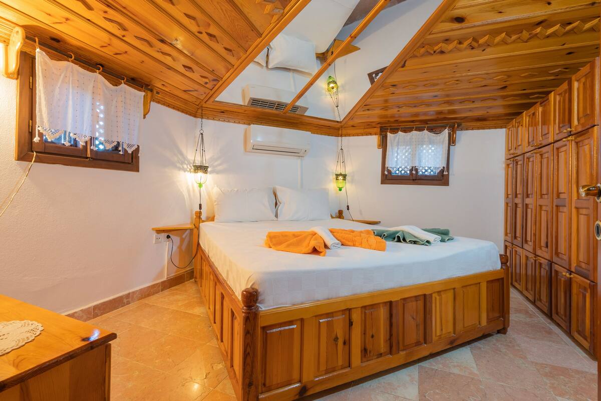 Een bed met houten frame, een wit matras, twee kussens en een opgevouwen handdoek. Twee nachtkastjes, waarvan één met een lamp en de ander met een stopcontact. De kamer heeft houten kastjes en een betegelde vloer.