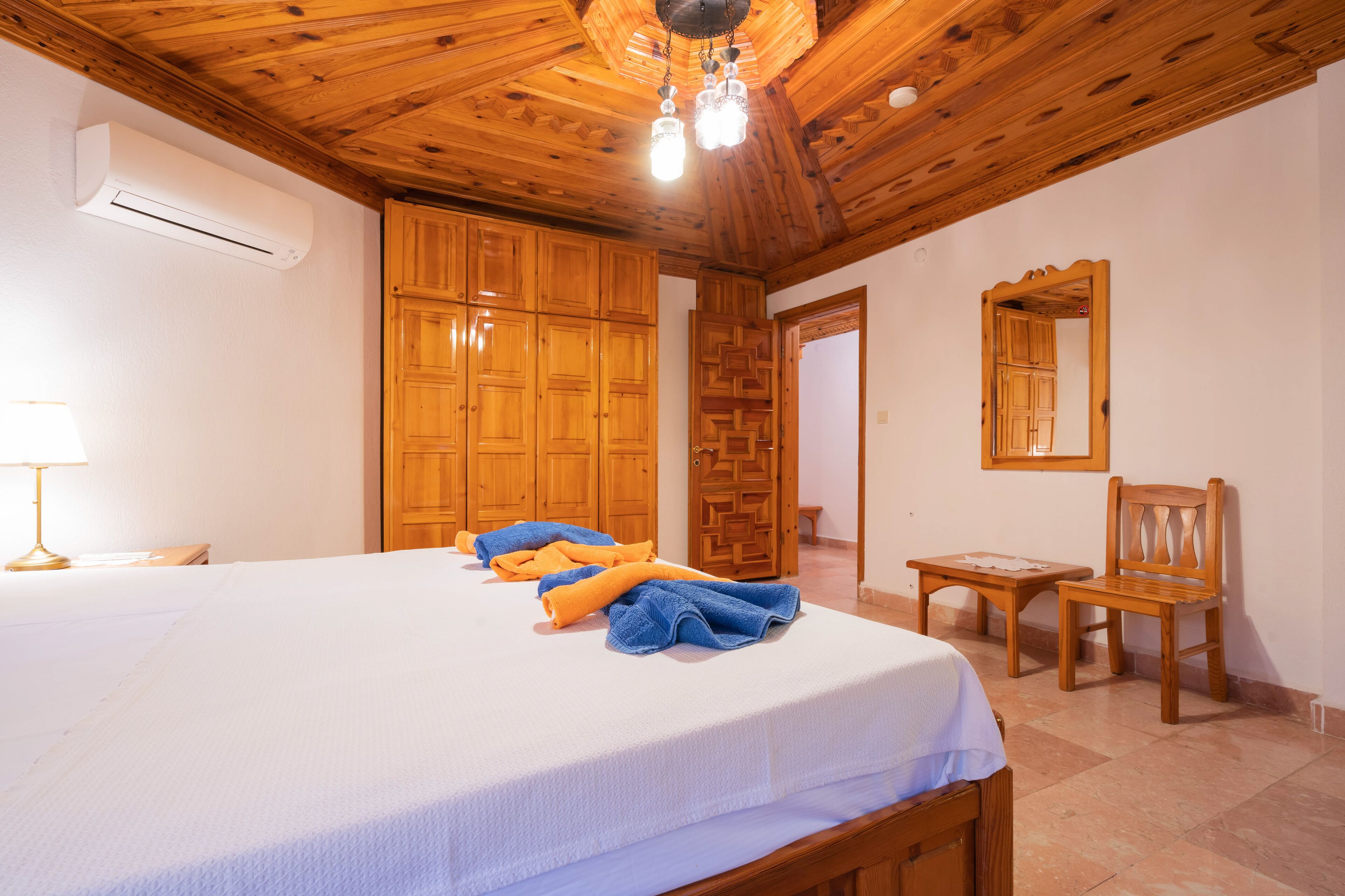 A bedroom with a large bed, wooden wardrobe, a wooden chair, and a mirror on the wall.