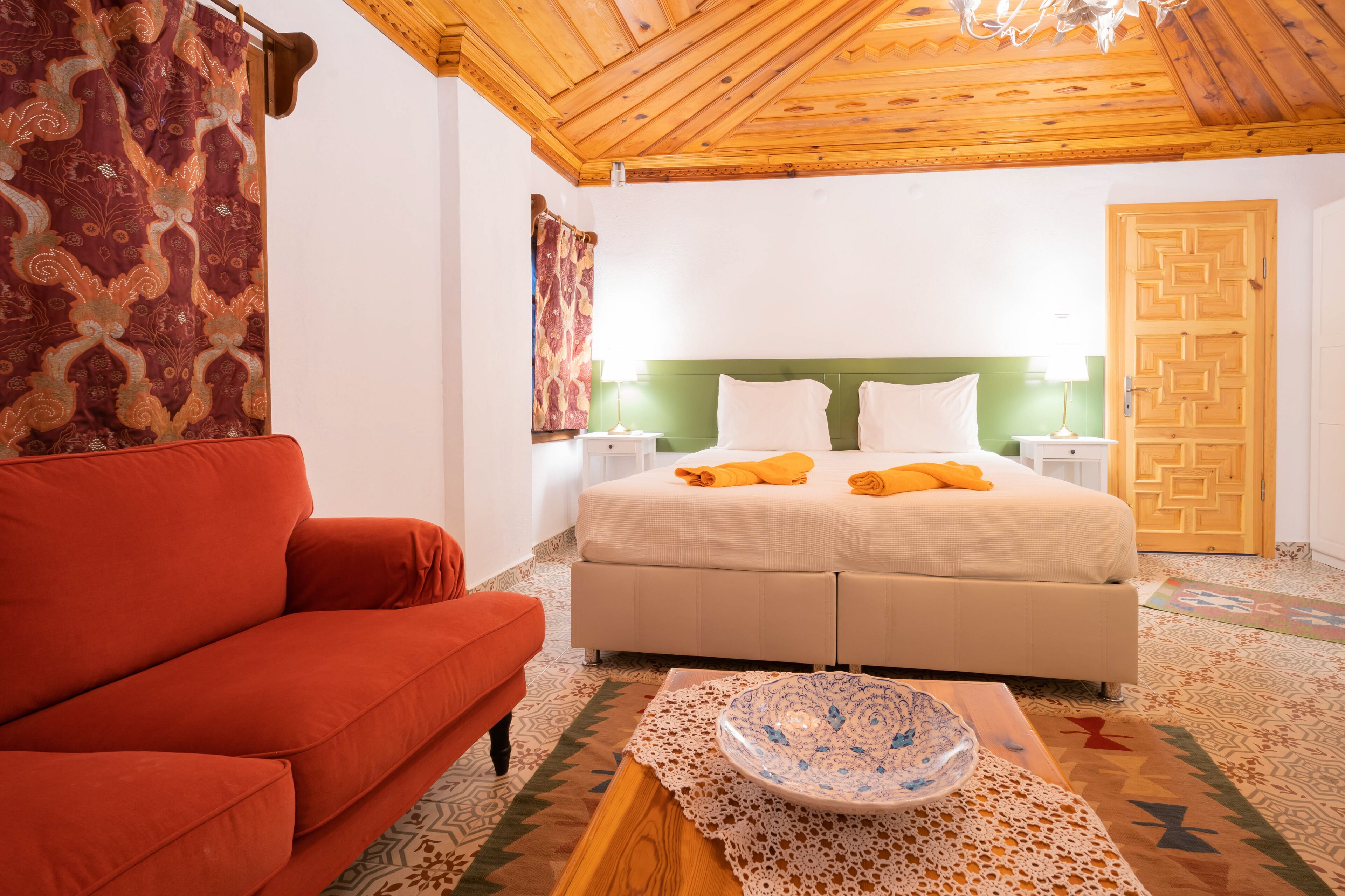 Un dormitorio con cama, sofá rojo, mesa de centro de madera con un cuenco, cortinas estampadas y techo de madera.