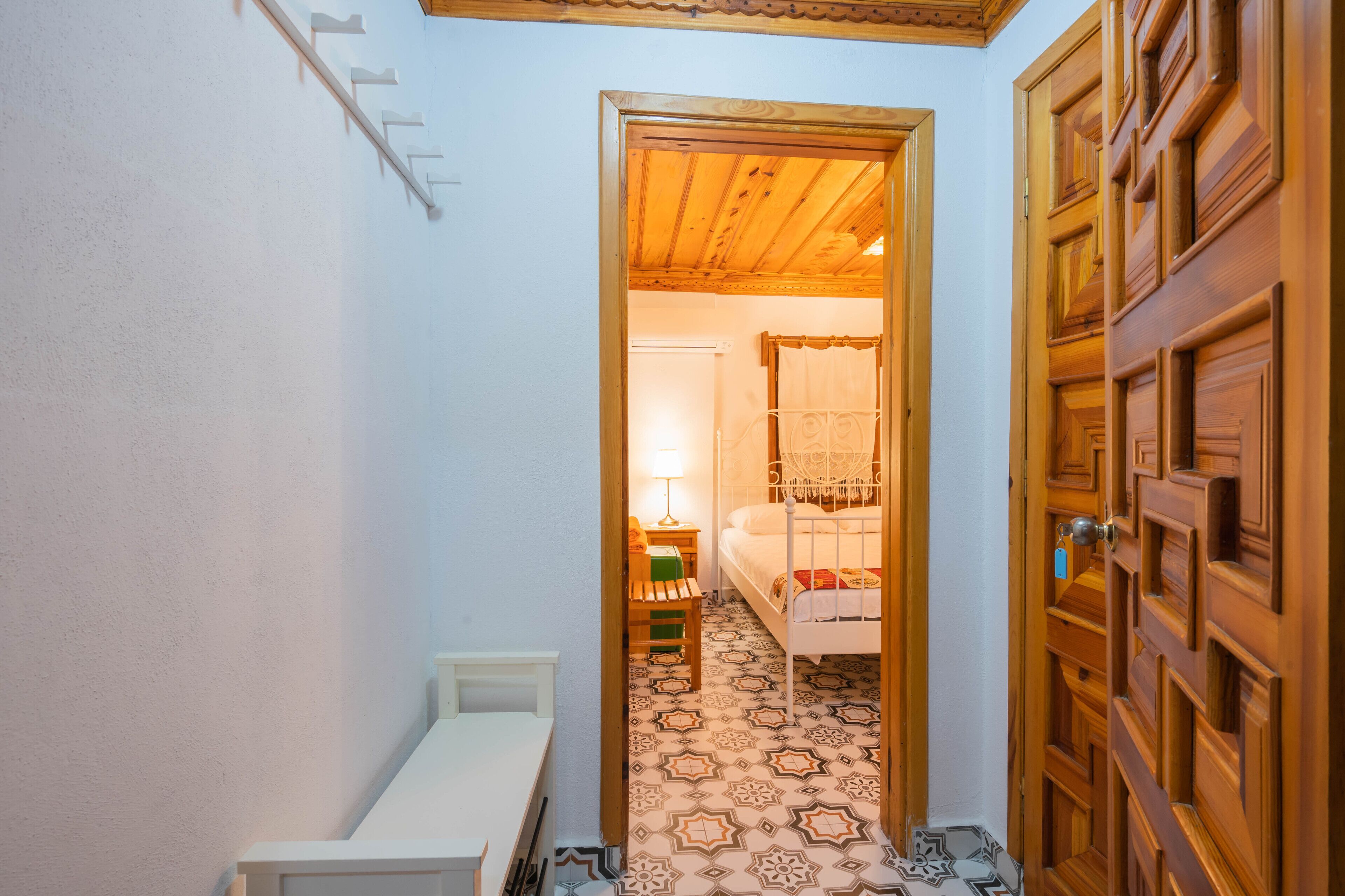 A narrow hallway with a wooden door on the right and a bed with a patterned floor in the background.
