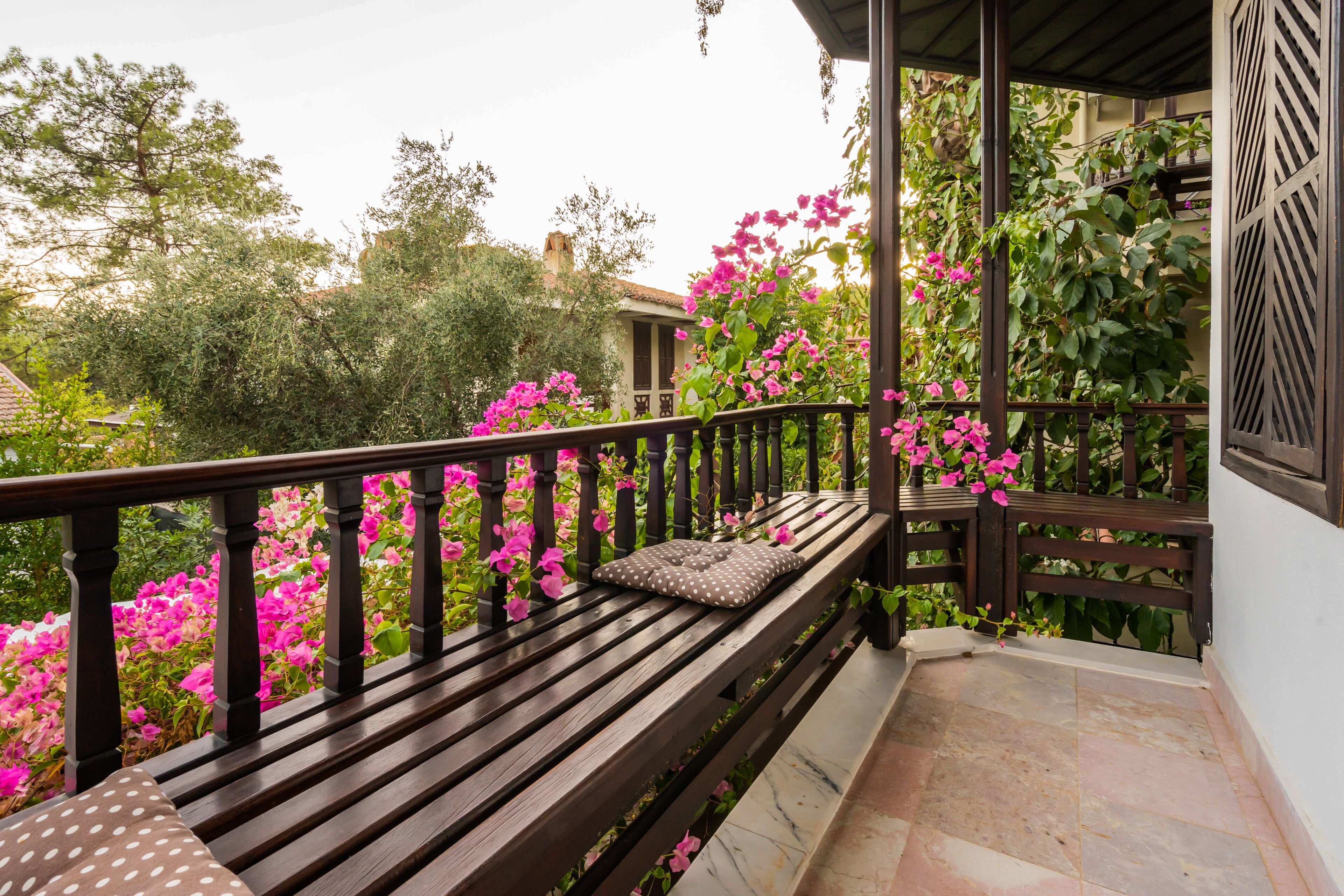 A wooden bench with a cushion on a balcony with a tiled floor, surrounded by pink flowers and greenery.