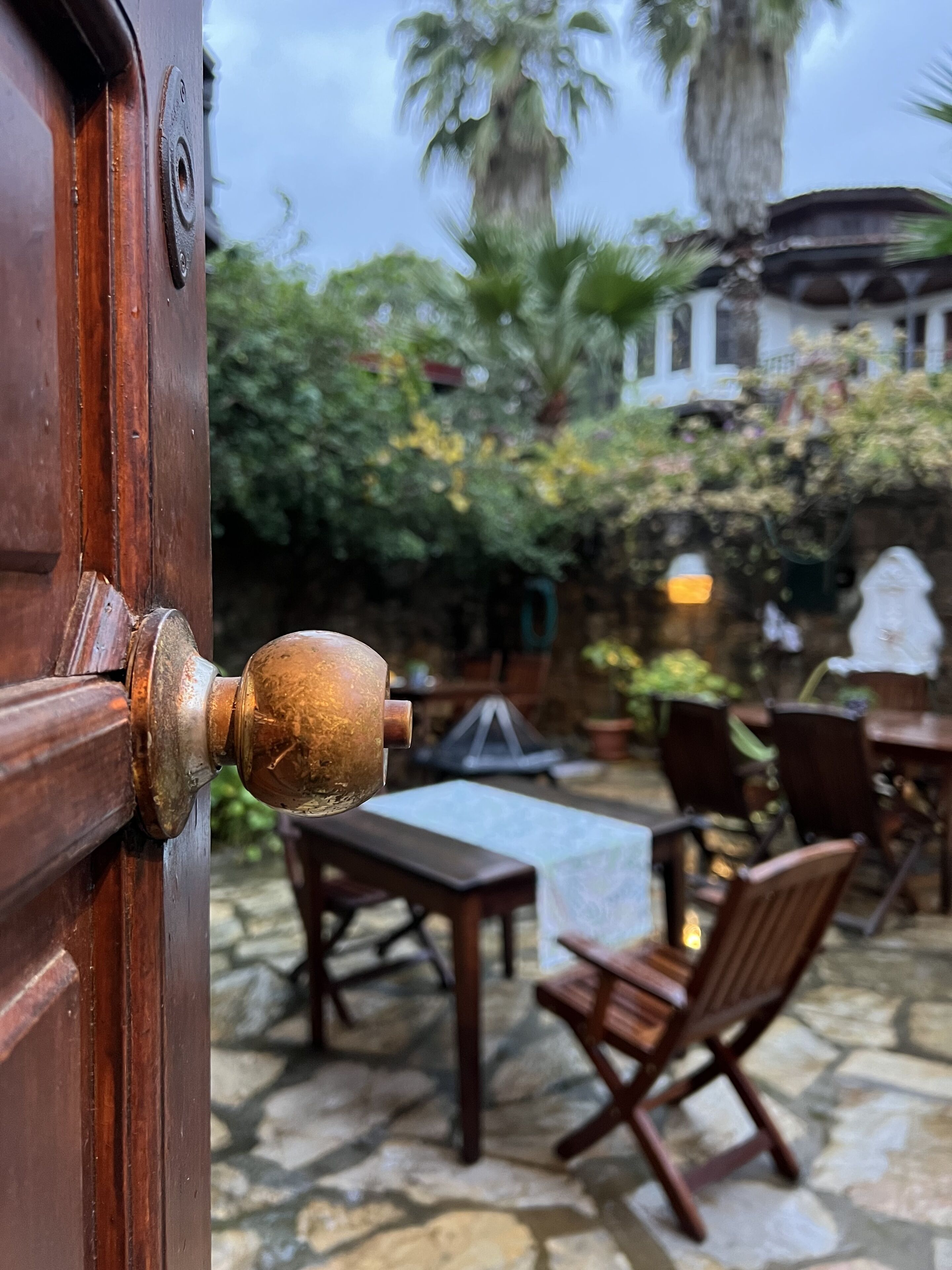 A wooden door with a brass doorknob, leading to an outdoor patio area with stone flooring, wooden furniture, and lush greenery.
