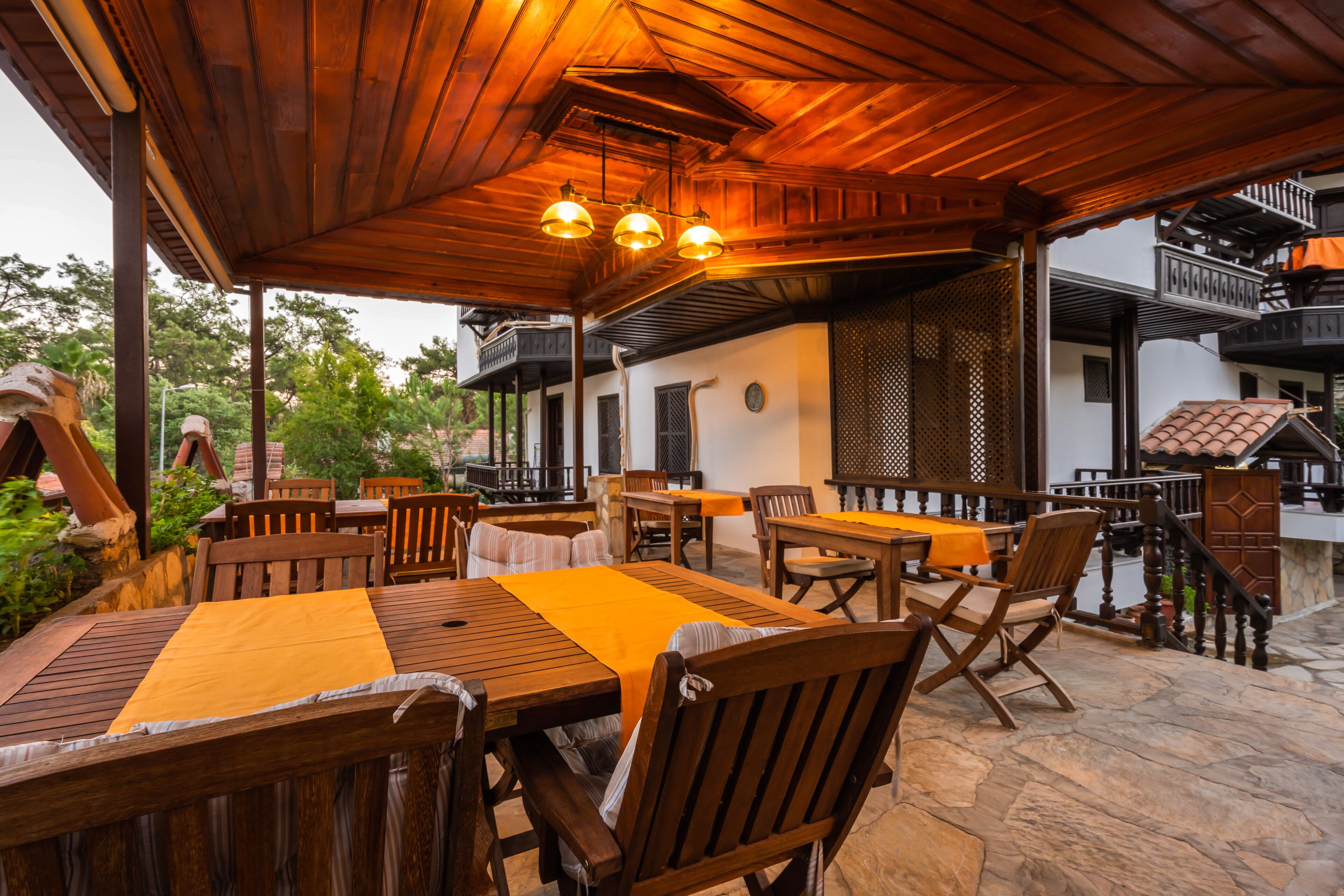 Un espace extérieur aménagé en terrasse, avec des tables et des chaises en bois, un coin salon abrité et un sol en pierre.