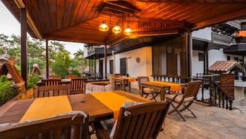 Un espace extérieur aménagé en terrasse, avec des tables et des chaises en bois, un coin salon abrité et un sol en pierre.