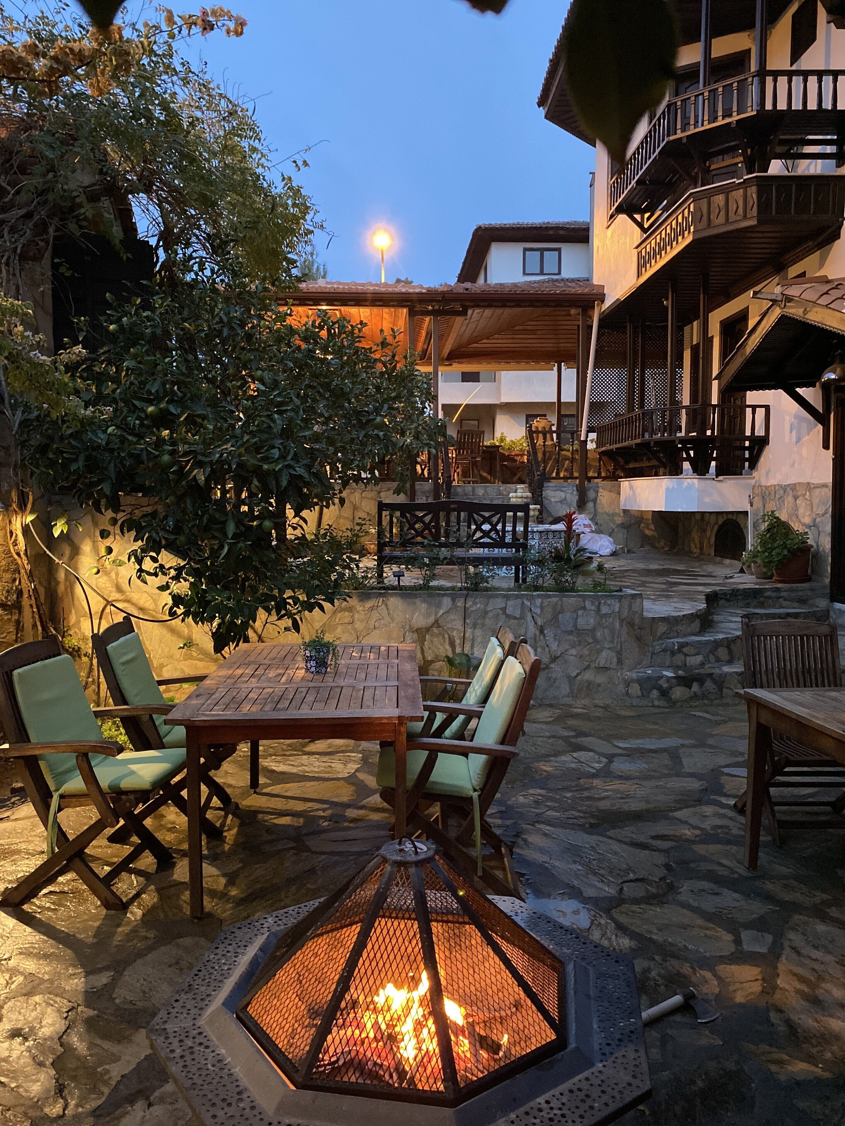 An outdoor patio with a fire pit, stone flooring, and wooden furniture.