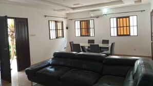 Een ruime woonkamer met een zwarte leren bank, een eettafel met stoelen en grote ramen voorzien van verticale zonwering.