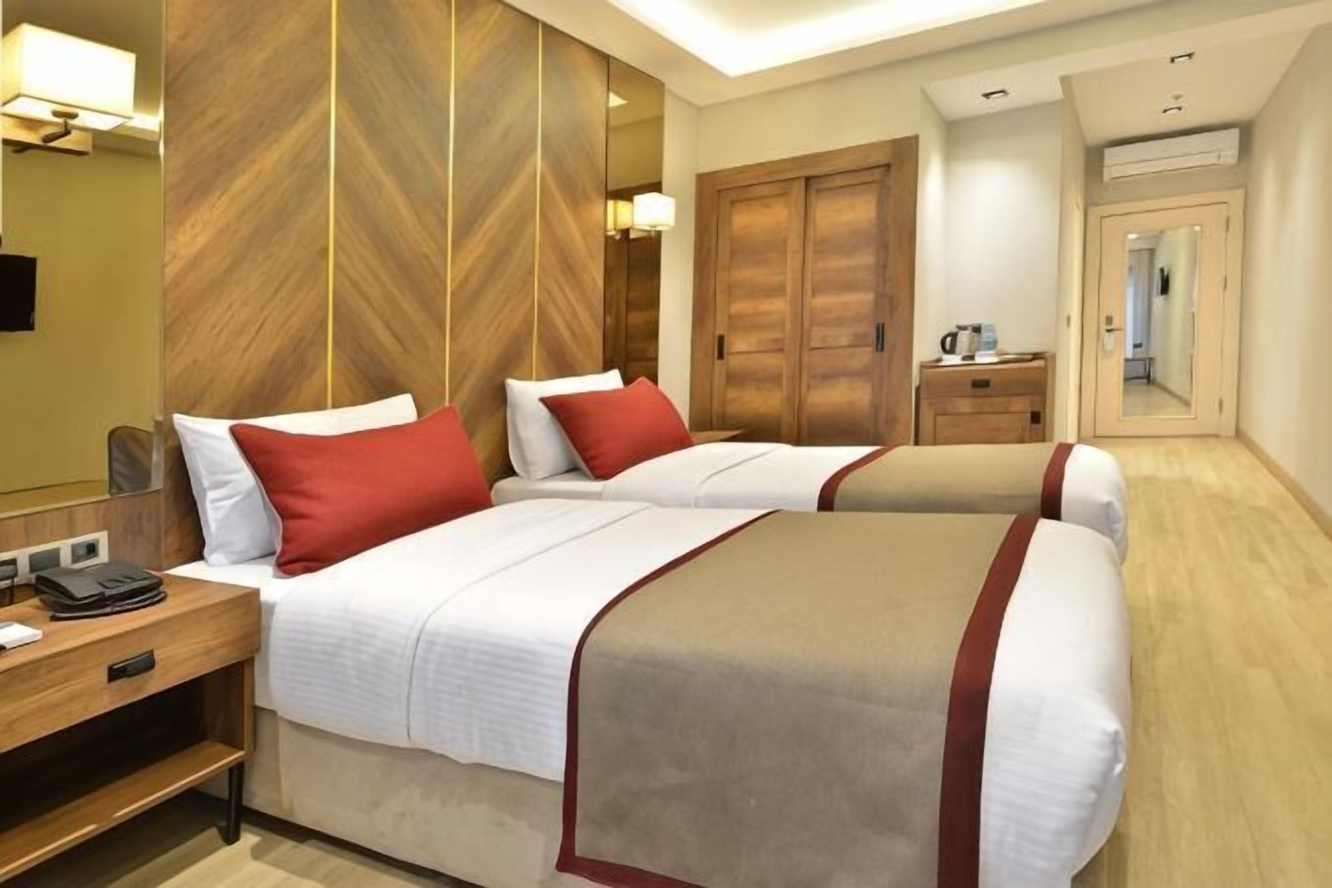 A hotel room with two beds, a wooden accent wall, a mirror, a desk with a phone, and a wardrobe.