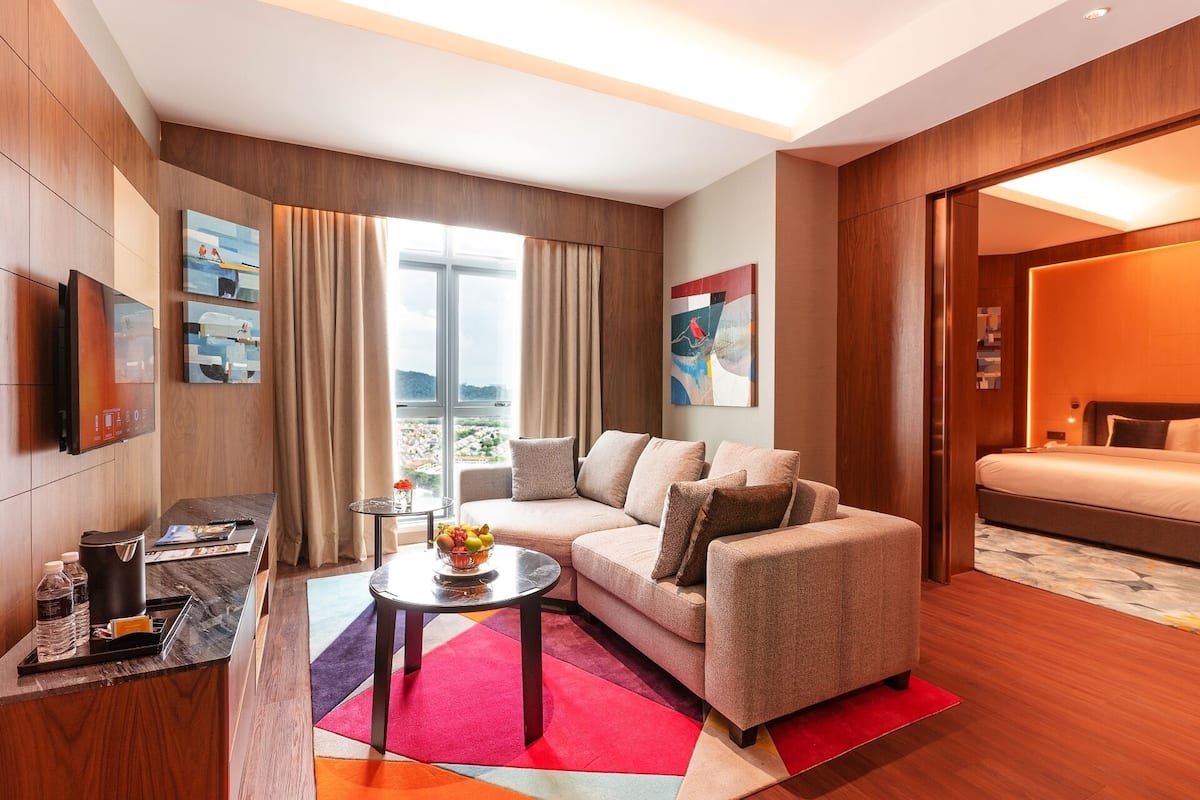 A modern hotel room with a sofa, a small table with a bowl of fruit, a TV, and a bed.