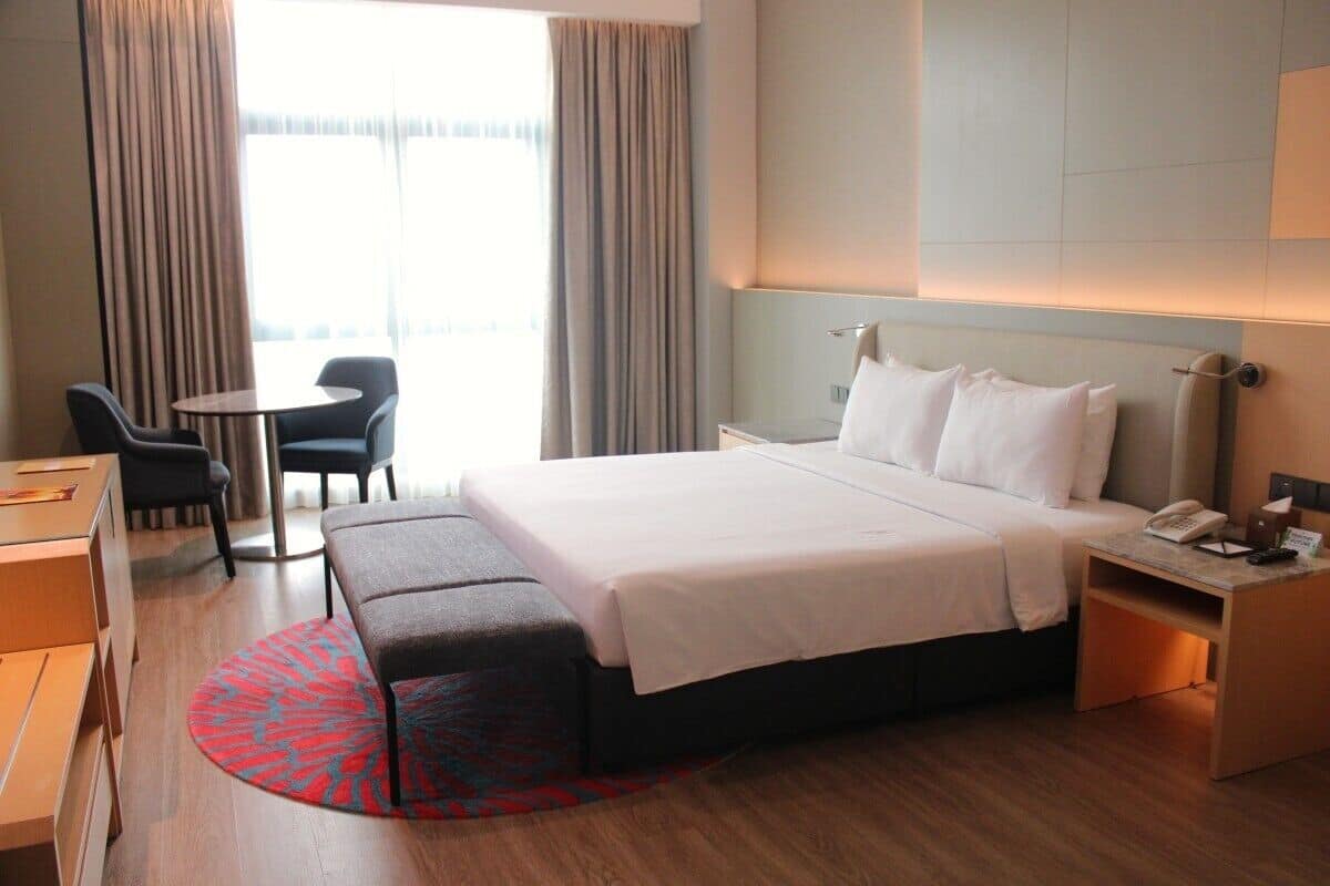 A hotel room with a bed, bedside table, chair, desk, and a view of the outdoors through large windows.