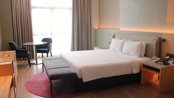 Habitación de hotel con cama, mesita de noche, silla, escritorio y vistas al exterior a través de amplias ventanas.