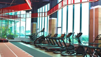 A gym with multiple treadmills aligned in rows, large windows offering a view of the city, and a red ceiling structure.
