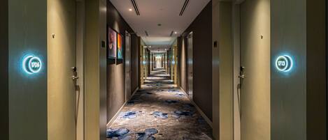 Corredor de hotel com portas em ambos os lados, carpete estampado e números de porta iluminados.