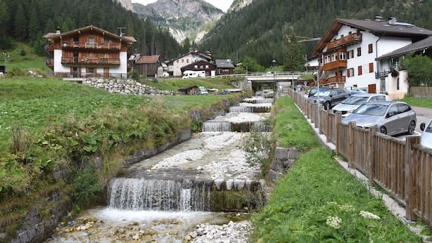 A mountain village with wooden houses, a small stream, and parked cars along the roadside. - Hotel Piz Boè (Mazzin)