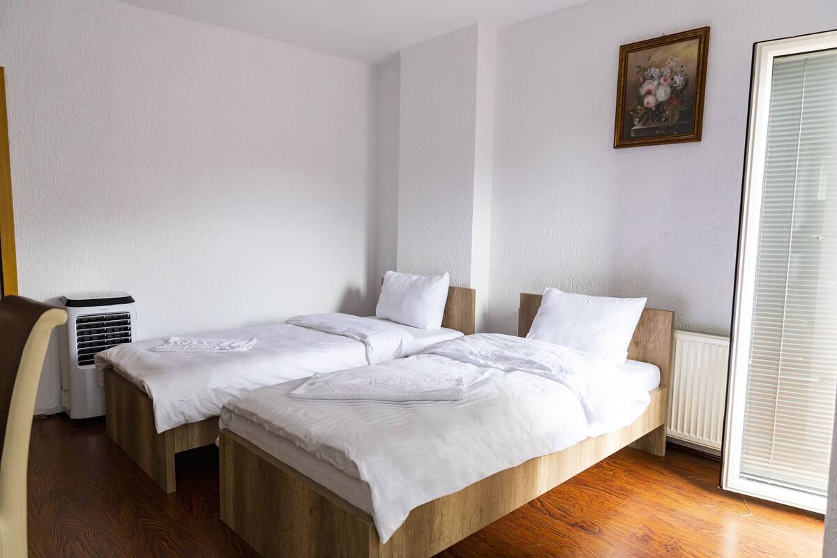 Ein Schlafzimmer mit zwei Holzbetten, weißer Bettwäsche, einem Heizkörper, einem Fenster mit Jalousien und einer Wand-Klimaanlage.
