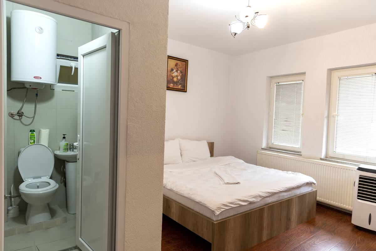 Ein Schlafzimmer mit einem Bett, einem Fenster mit Jalousien, einem Heizkörper und einem Duschbereich mit Toilette.
