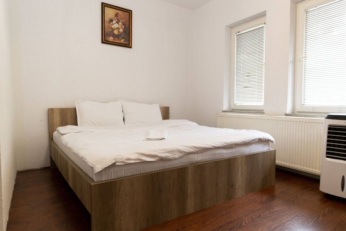 Ein Schlafzimmer mit einem Holzbett, weißer Bettwäsche, einem Heizkörper und einem Fenster mit Jalousien.