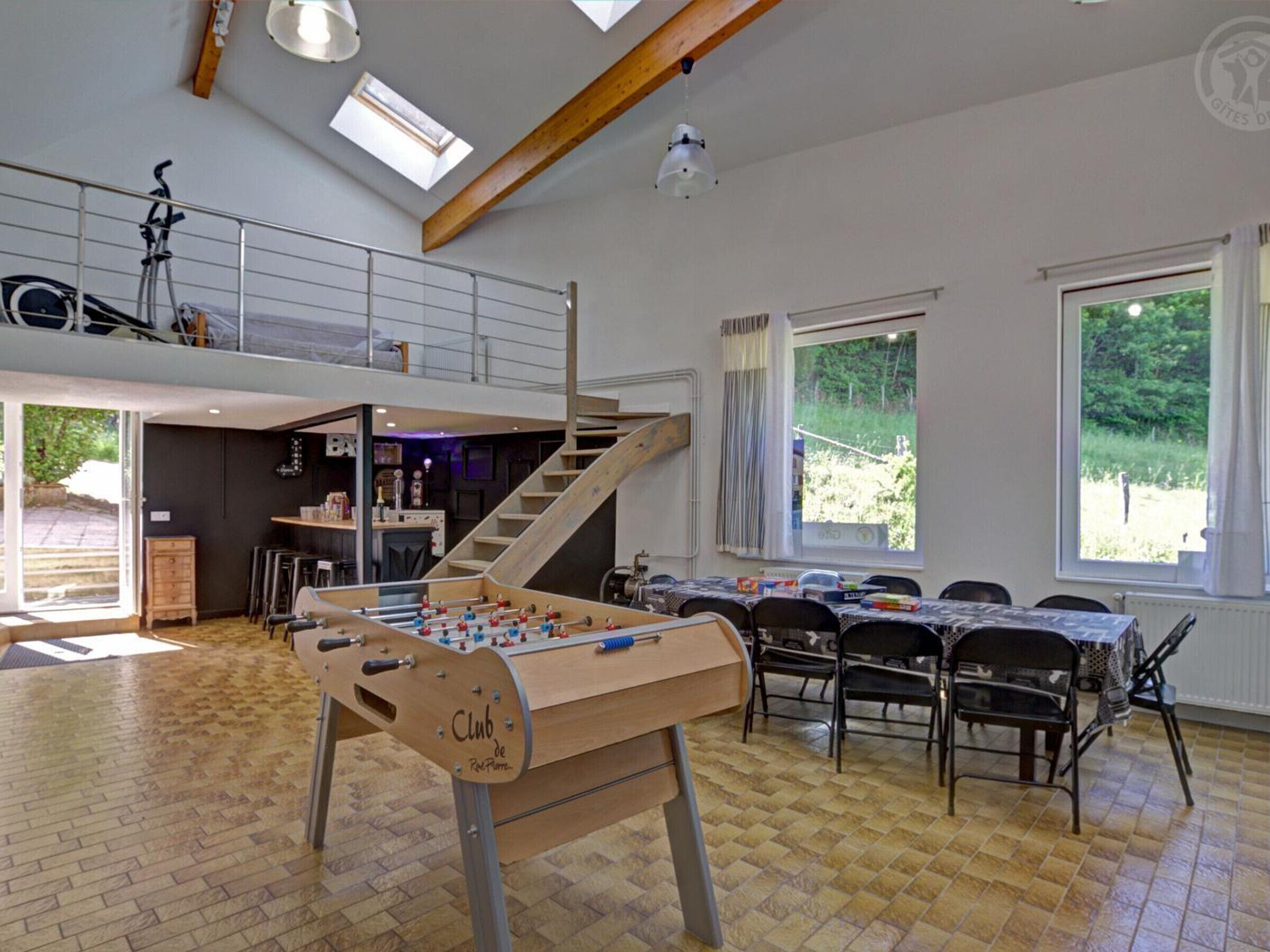 A spacious room with a foosball table, a dining area with tables and chairs, a staircase, and large windows offering views of greenery.