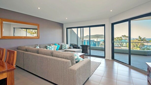A modern living room with a grey sofa, a wooden coffee table, and a large window offering a view of a balcony and the sea.