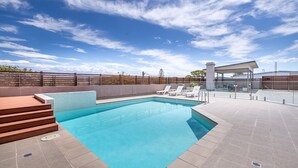 A rectangular swimming pool with a wooden deck and steps leading into it, surrounded by a fenced area with lounge chairs and a covered seating area.