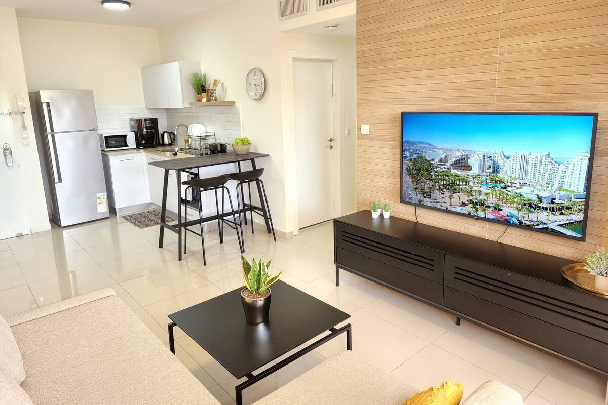 A modern kitchen with a refrigerator, microwave, and sink. There is a dining area with a table and chairs. A television is mounted on the wall, and a plant is on the coffee table.