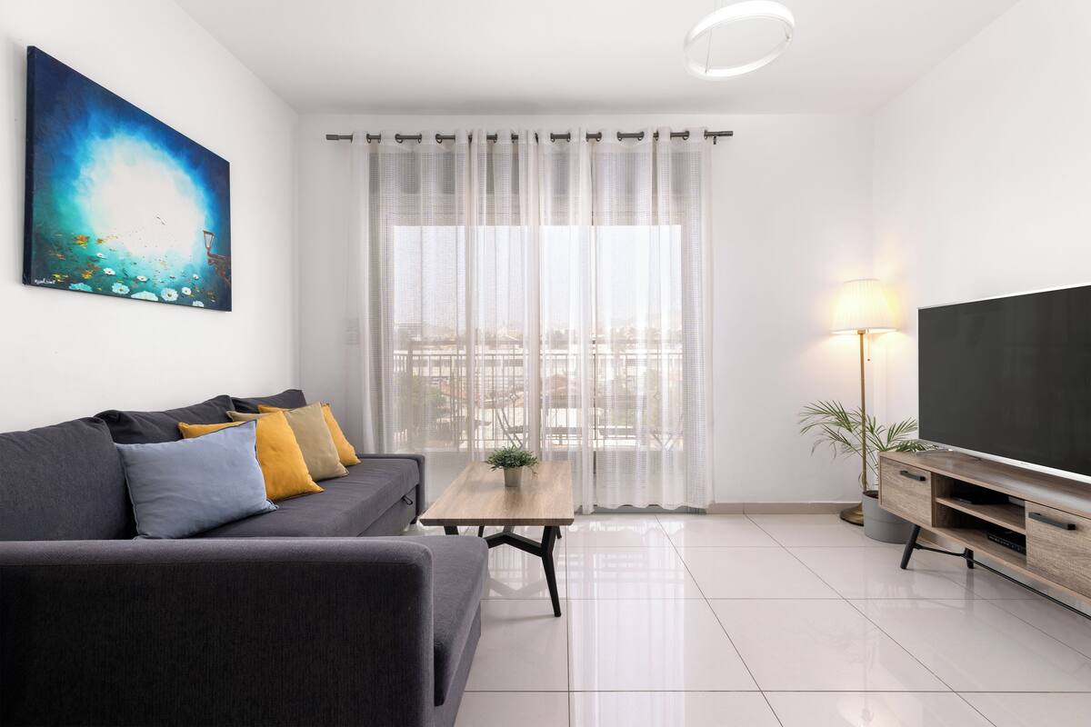 A modern living room with a sofa, a coffee table, a TV, and a large window with sheer curtains.