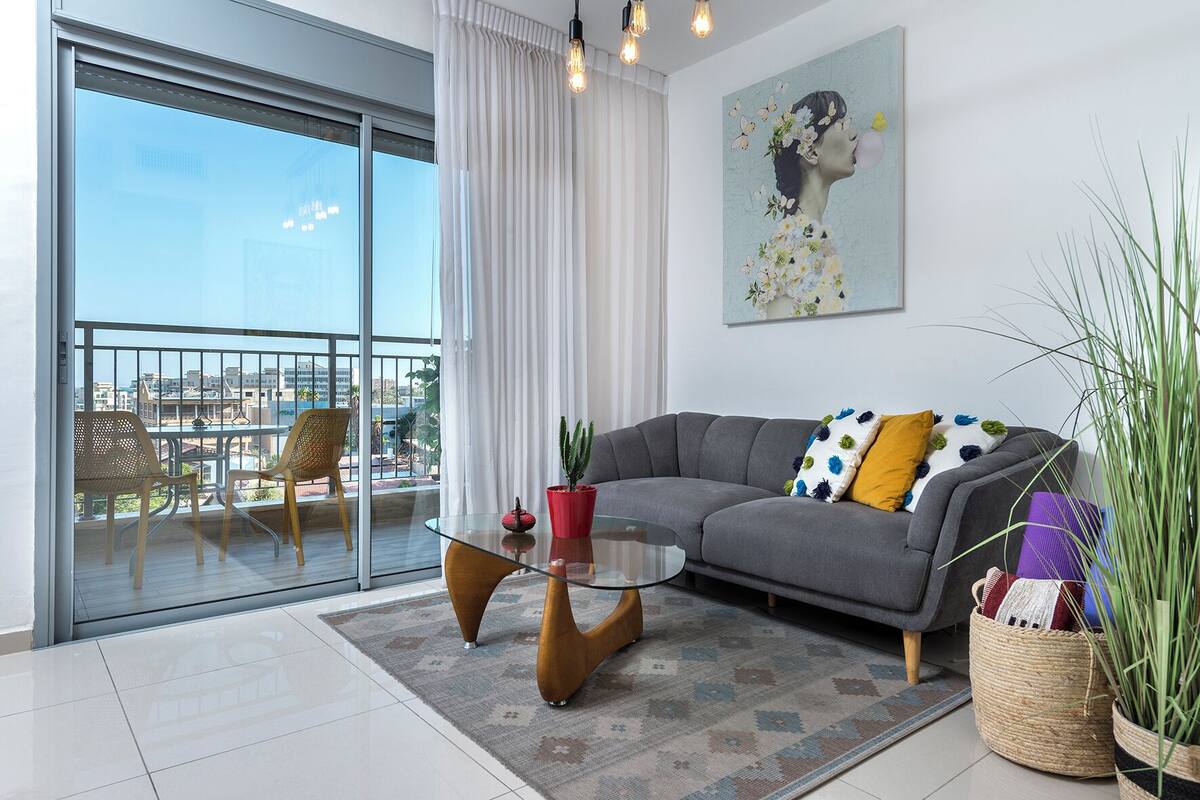 A modern living room with a grey sofa, a glass coffee table, and a view of the city through a sliding glass door.