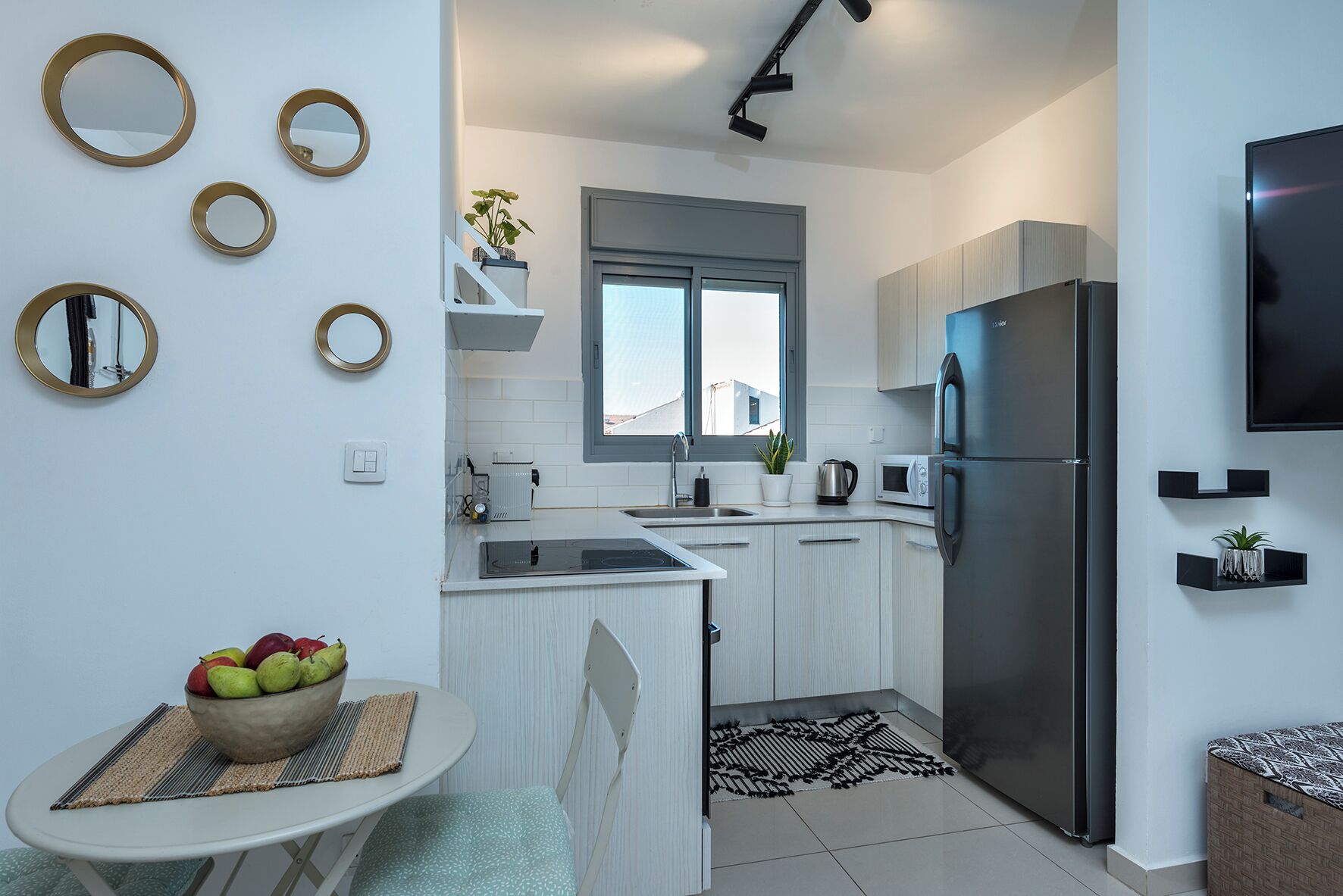 Una cucina moderna con mobili bianchi, un frigorifero in acciaio inossidabile, un forno a microonde e un lavello. Si notano specchi a parete, un piccolo tavolo con una ciotola di frutta e una finestra sopra il lavello.
