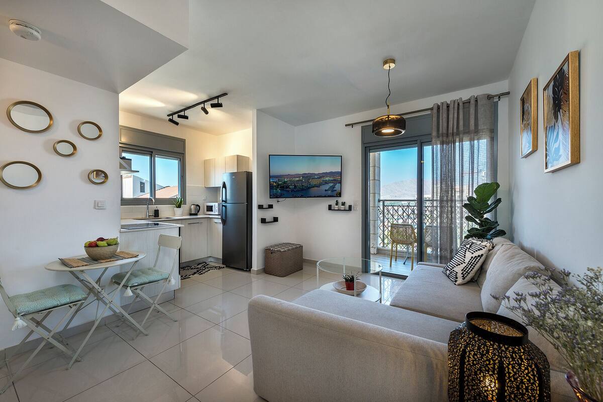 A modern apartment with a kitchen, dining area, and living room. It features a balcony with a view, a sofa, and a dining table.