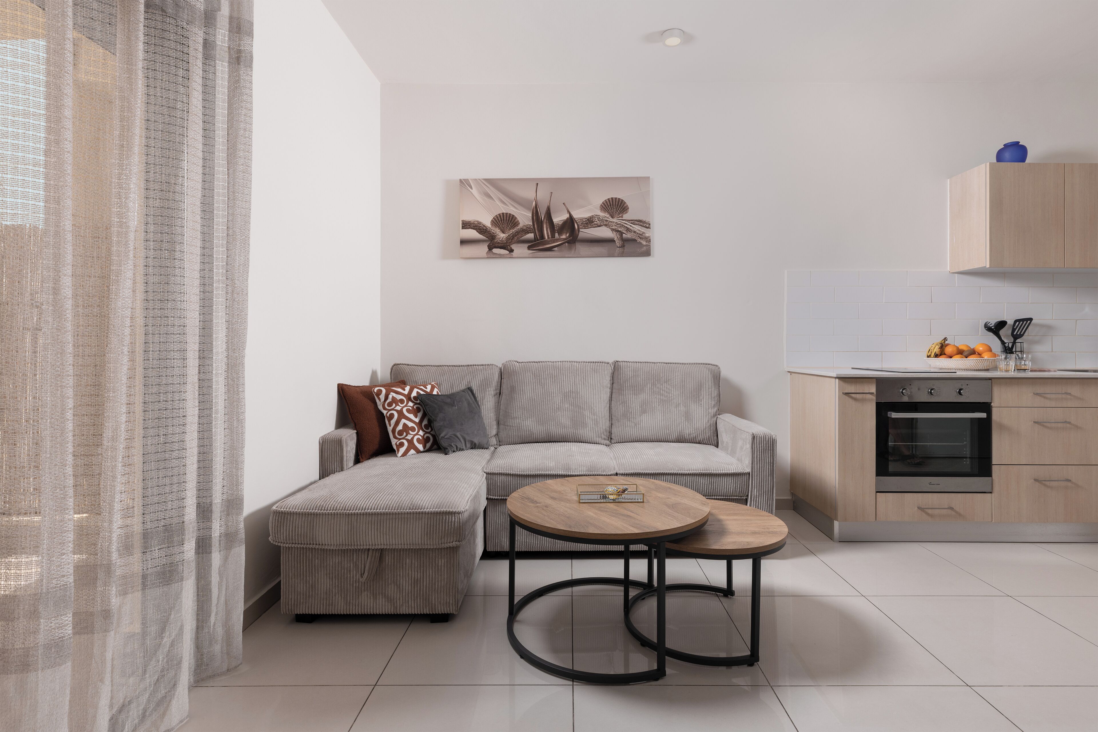 A modern living room with a sofa, ottoman, and round coffee table. There is a kitchen area with cabinets and a stove.
