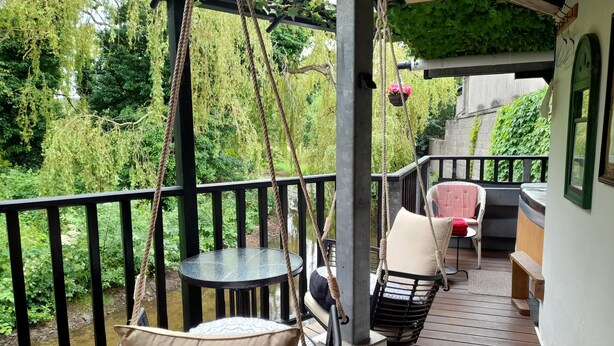 A wooden deck with a swing chair and a table, overlooking a garden with a stream.