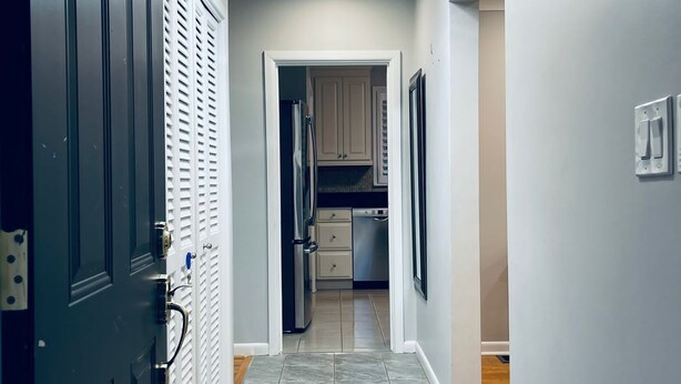 A hallway with a dark door, a white wall, and a kitchen area visible through an open door. - "The Cottage at Spruce" Tranquil, Cozy and Rustic (Burlington)