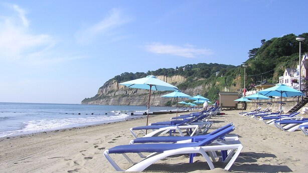 A beach with rows of sun loungers and umbrellas, a cliff in the background, and a building with stairs leading up to it.