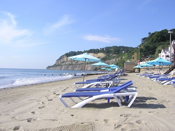 A beach with rows of sun loungers and umbrellas, a cliff in the background, and a building with stairs leading up to it.