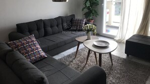 A modern living room with a grey sofa, a small round table with a vase of flowers, and a large window with sheer curtains. - Superb Mont Carlo Hamam Sousse vacation apartment (Hammam Sousse)