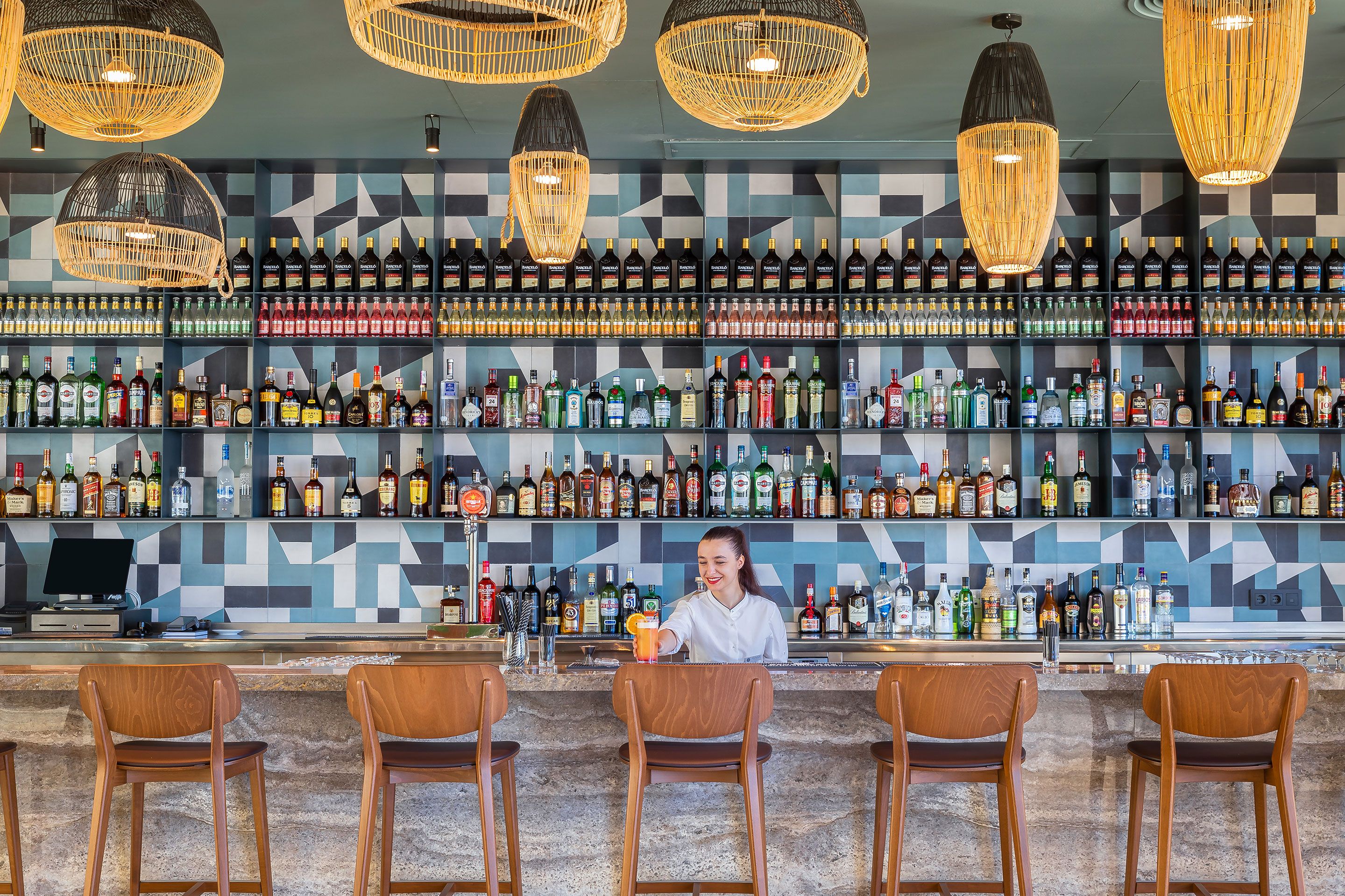 A bar with multiple liquor bottles on shelves, a bartender preparing a drink, and three wooden bar stools.