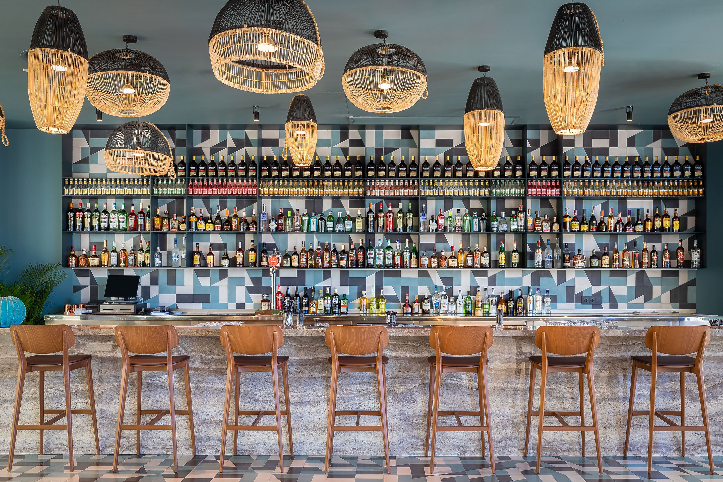 Uma área de bar com várias garrafas de bebidas, banquetas de madeira e luminárias de vime suspensas.