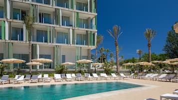 Area piscina di un hotel con lettini e ombrelloni, circondata da palme e un edificio a più piani con balconi.