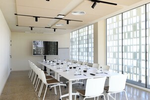 Un salón de conferencias con una larga mesa, sillas blancas, una pizarra y un ventanal grande.