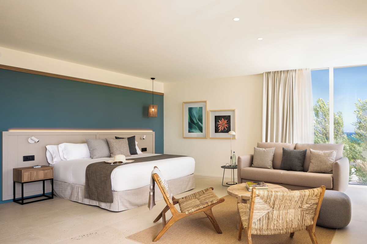 A modern hotel room with a bed, a sofa, a chair, a small table, and artwork on the walls.