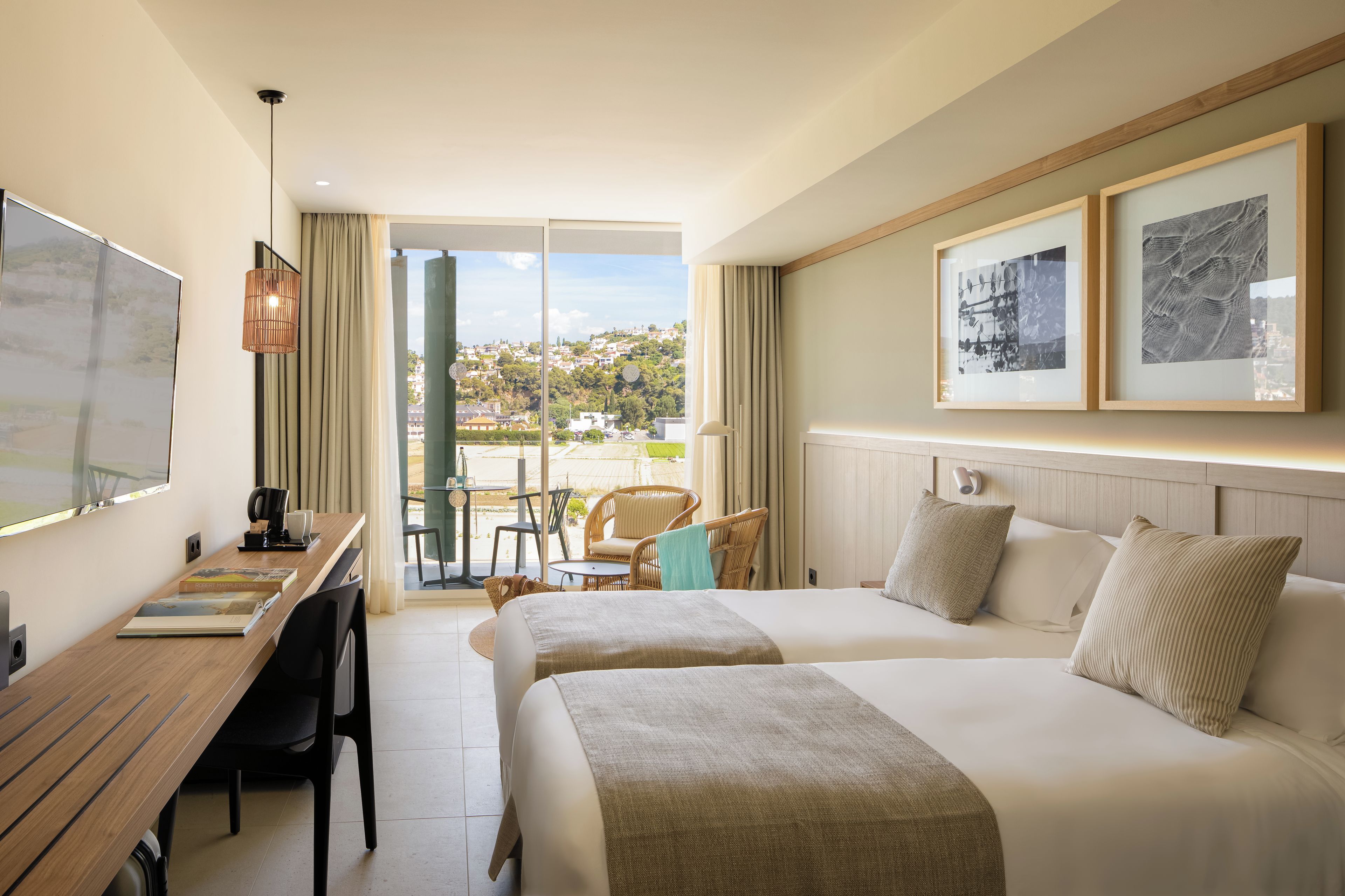 A hotel room with two beds, a desk with a chair, a table with chairs, and a view of the city.