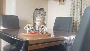 Zona pranzo con tavolo in legno, due sedie nere, tavolino con una tazza e snack, cesto in rete nera e televisore a parete.