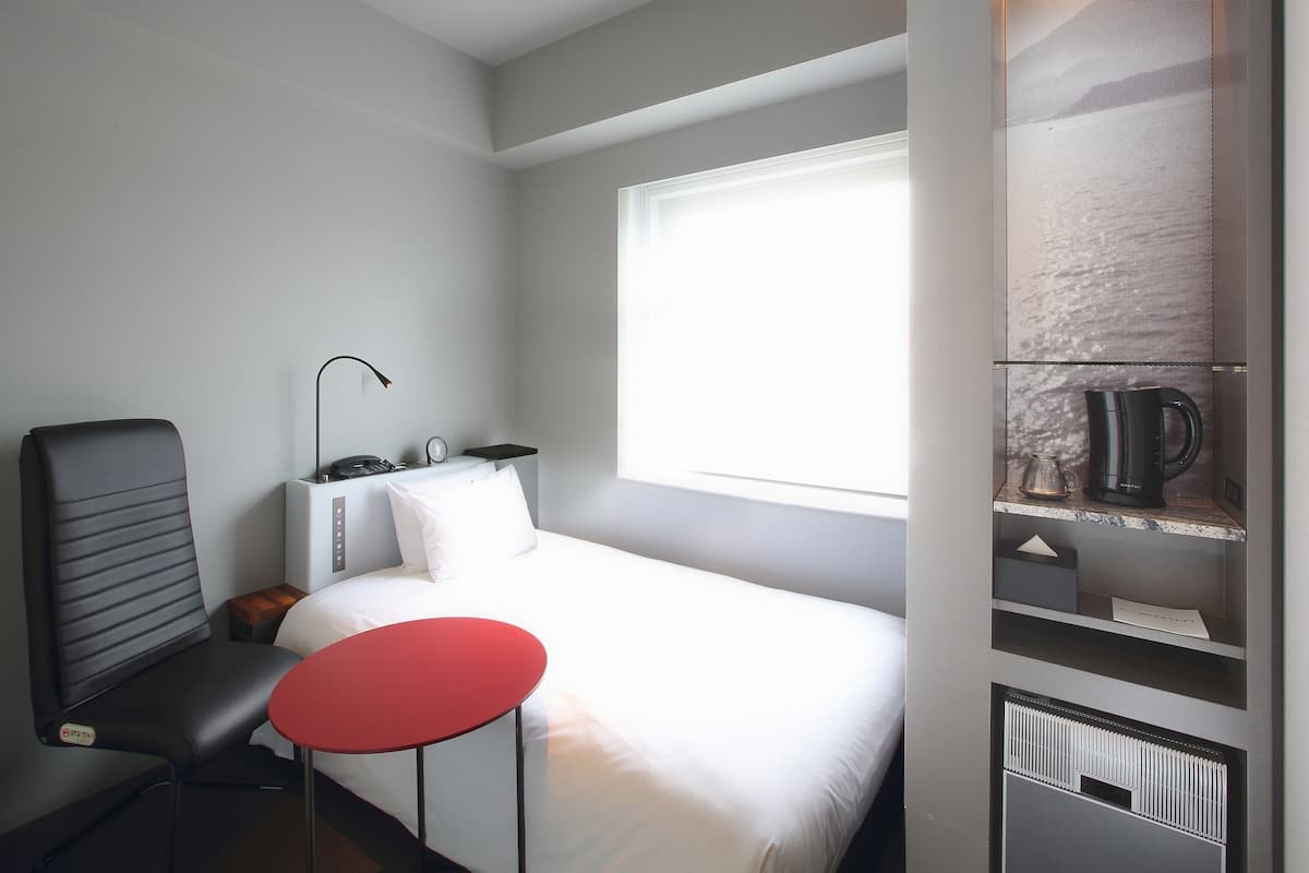 A hotel room with a bed, a red table, a chair, a desk with a lamp, and a window.